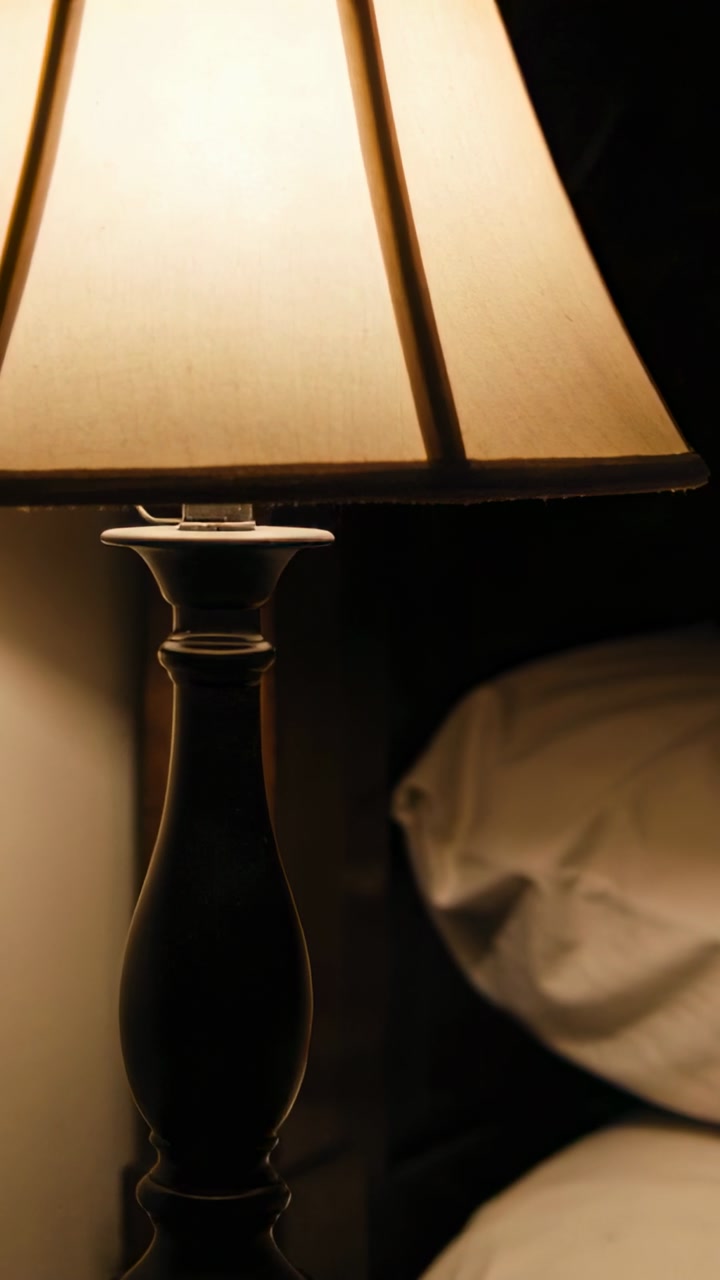 Vertical video: Forearm shifting, bare adult hand reaching for table lamp base on nightstand pillow