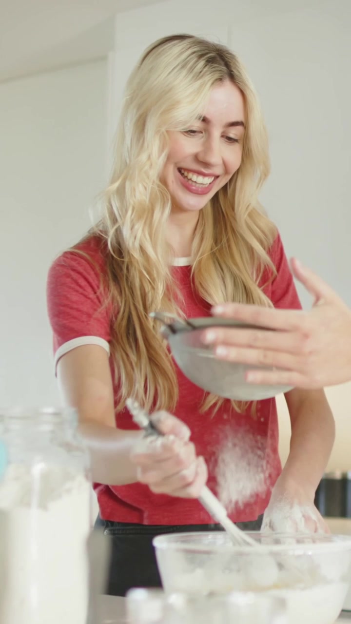 Video verticale: Donna con maglietta rossa che mescola l'impasto per cuocere in cucina, setaccia la farina