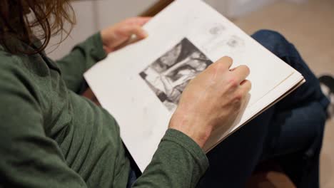 por encima del hombro, una foto de un artista con el pelo largo y castaño con una camisa verde de manga larga sentada dibujando en un cuaderno de bocetos