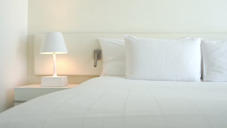 En-Una-Habitación-De-Hotel-Blanca,-Hay-Una-Lámpara-Blanca-Sobre-Una-Mesita-De-Noche-Blanca-Que-Ilumina-Una-Cabecera-Y-La-Ropa-Blanca-Y-Fresca-De-Una-Cama-Perfectamente-Hecha