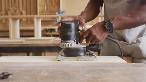 mid section of african american male carpenter using an electric grinder to grind wooden plank