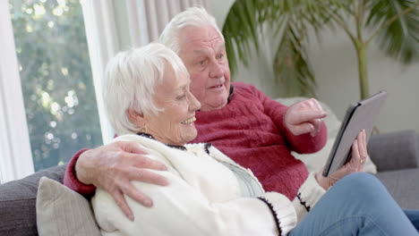 Feliz-Pareja-Caucásica-Mayor-Sentada-En-Un-Sofá-Usando-Una-Tableta-En-Casa,-Cámara-Lenta