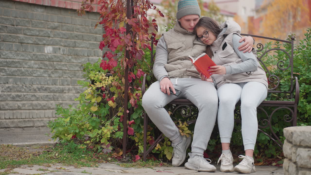 Bonding moment between young couple seated outdoor on garden bench with husband gently patting her hand and kissing her head while reading together surrounded by autumn foliage