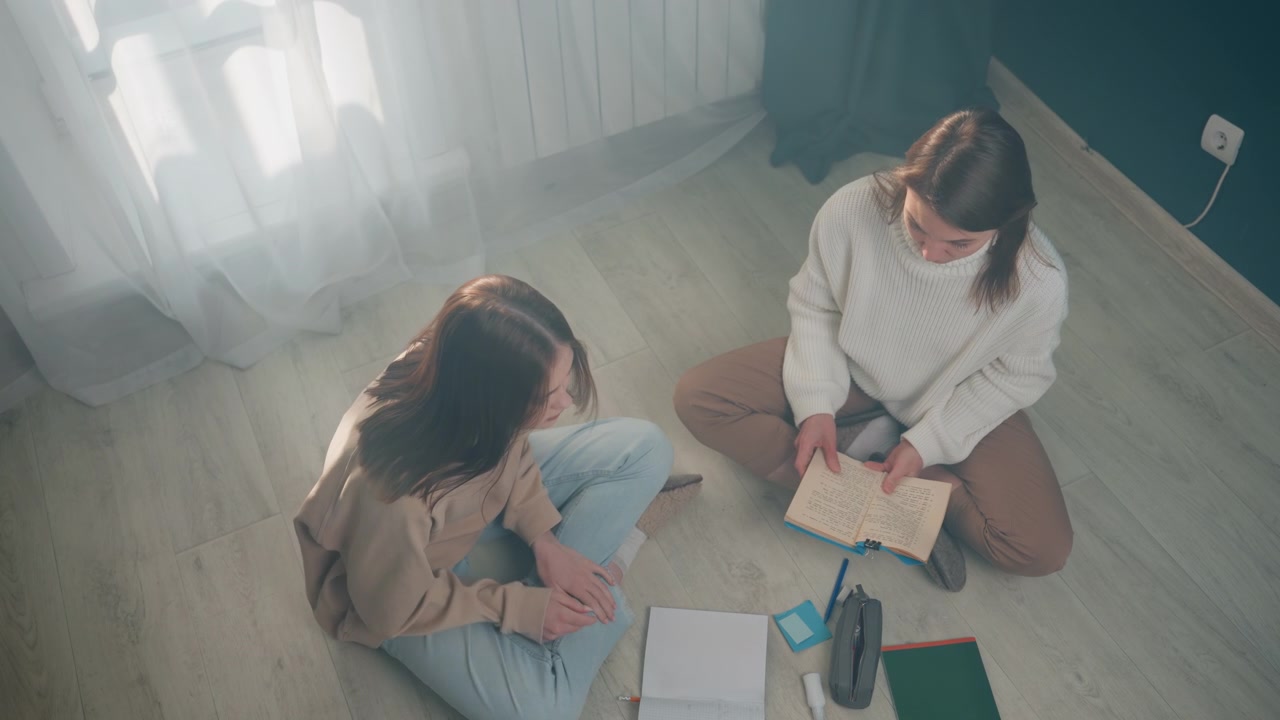 自宅で、床に座ってノートとタブレットを使い文法を練習する2人の女性。暖かな日差しが差し込む中、カジュアルなセーターを着てリラックスした家庭教師のセッションが行われている。熱心な学生がコーチングを受け、穏やかな訂正がなされる。