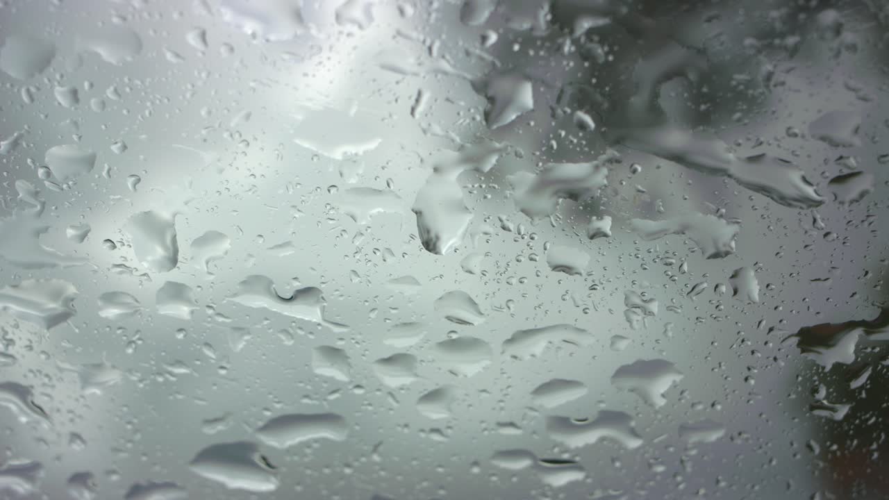 Detailed view of rain drops cascading down a car window during a heavy storm. The dynamic, water-streaked glass creates an abstract backdrop for the dramatic, moody atmosphere
