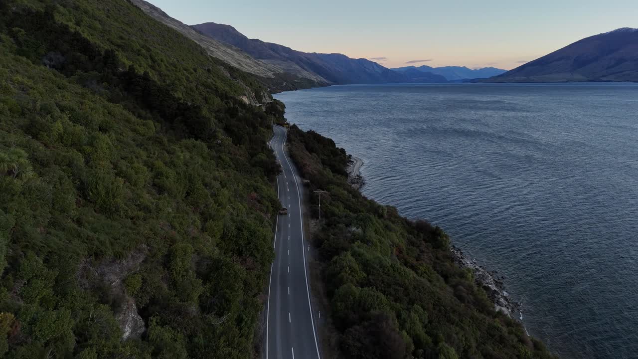 Aerial tracking shot of car on coastal road beside lake hawea in New Zealand. Peaceful sunrise behind mountains and hills of South Island