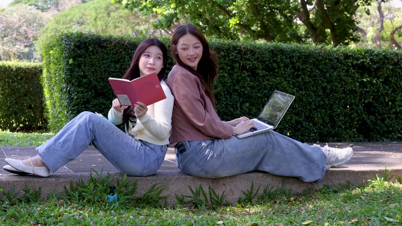 屋外で背中合わせに座った2人のアジア系の女子学生が、ノートパソコンとノートを使って勉強している。
