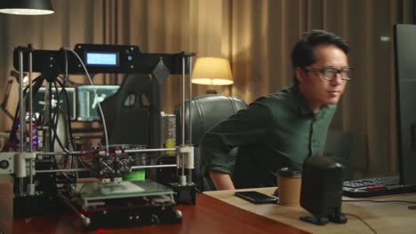 asian man start works on personal computer and 3d printer in home office, he works at night