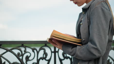 dama sosteniendo un libro abierto en su mano con la mano derecha en las páginas mientras la mano izquierda apoya debajo, el viento sopla suavemente las páginas, con un fondo borroso con cuerpo de agua y vegetación en la distancia
