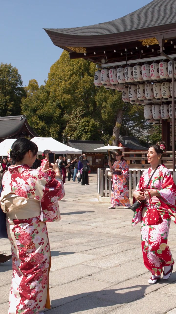 Turisti in kimono colorati camminano attraverso un cortile di tempio soleggiato con lanterne di carta appese.