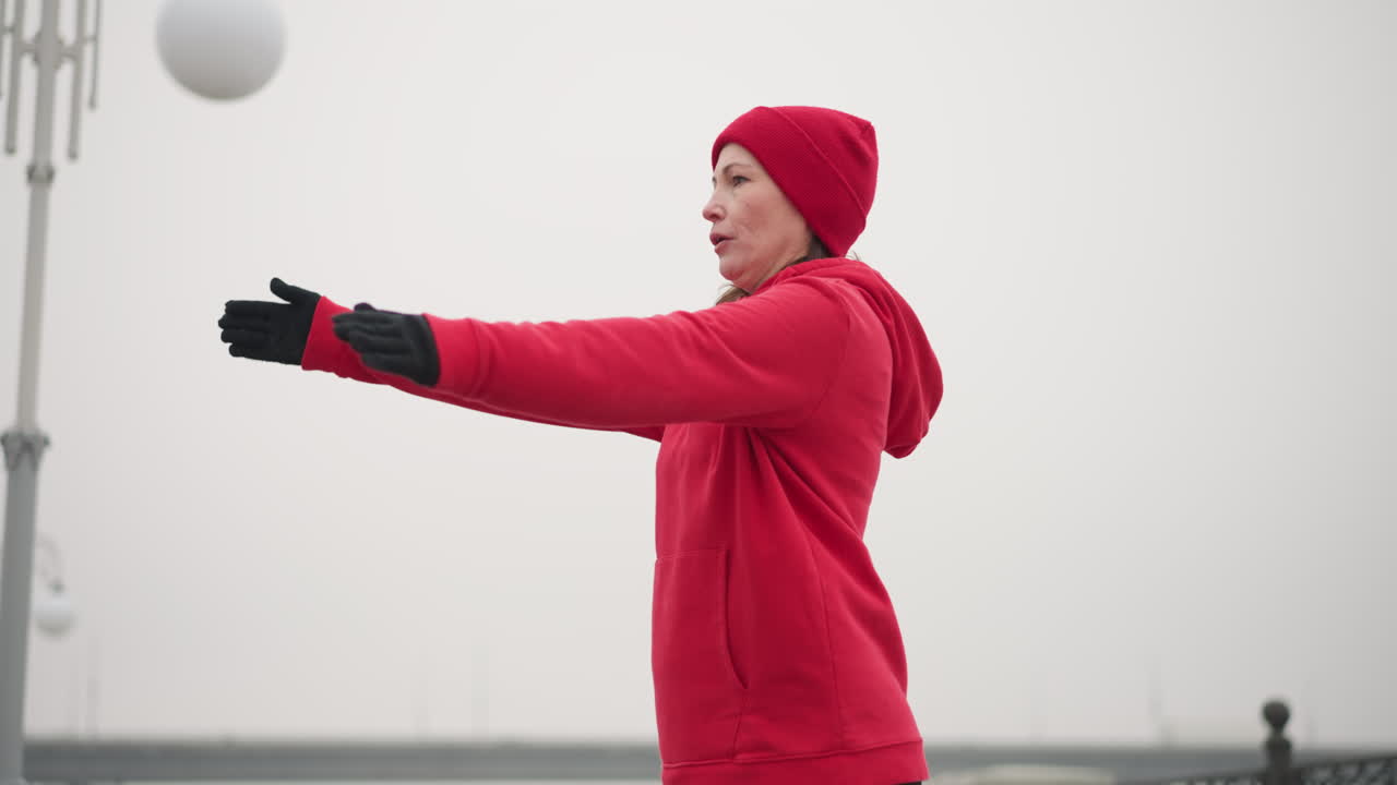 은 후디와 장갑을 입은 여성이 야외에서 운동을 하는 동안 안개가 많은 겨울 환경에서 빛의 기둥과 최소한의 도시 특징을 배경으로합니다.