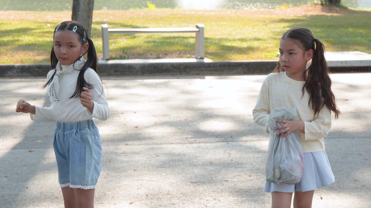 Two young girls in casual clothing perform dance moves in a sunny outdoor park setting