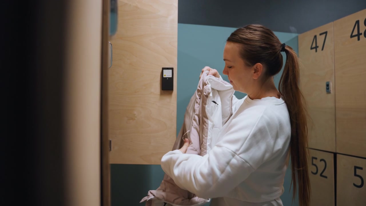 Locker room white woman adjusting puffer jacket before badminton practice, numbered wooden lockers, teal wall, soft overhead light, focused expression as she zips coat and checks locker space in quiet