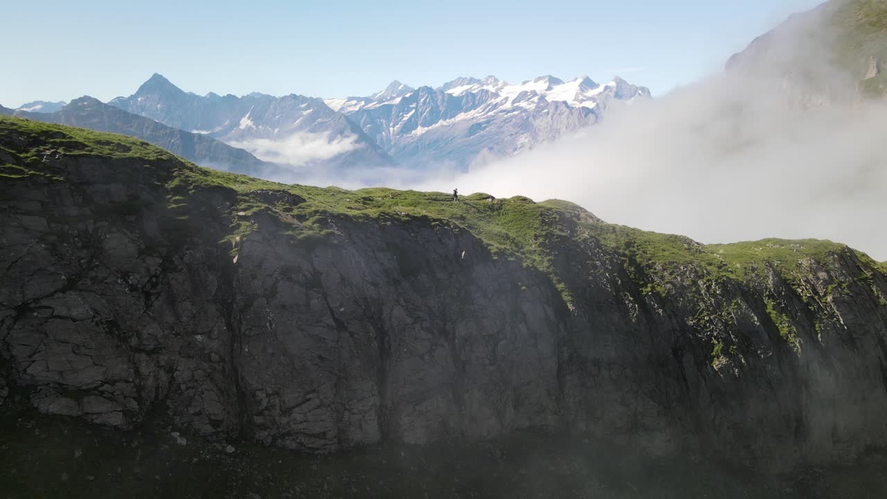 Einzelwanderer geht einen schmalen alpinen Grat entlang, mit dramatischen Berggipfeln und Wolken unter sich. Kinoreife Luftaufnahme, die Abenteuer, Ausgesetztheit und Einsamkeit in den Schweizer Alpen zeigt