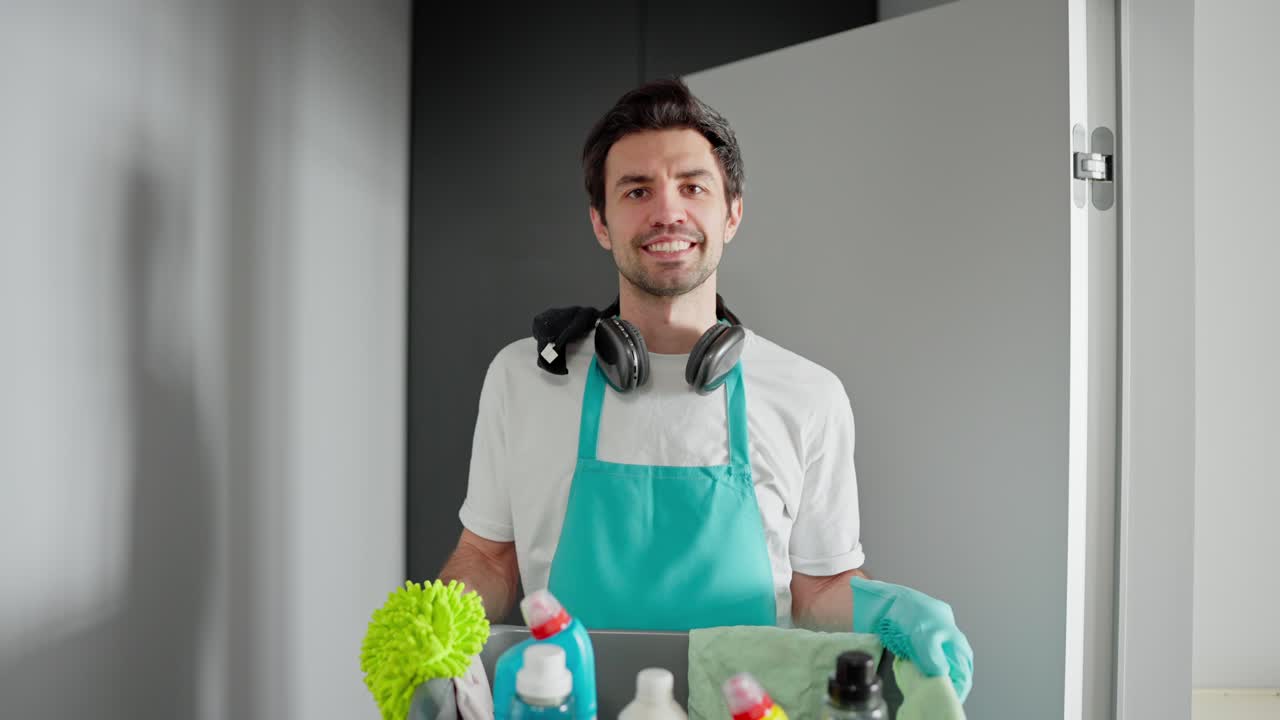 retrato de un limpiador masculino confiado en una camiseta blanca y un delantal azul caminando y llevando un cuenco de plástico gris con herramientas de limpieza y detergentes en un apartamento moderno