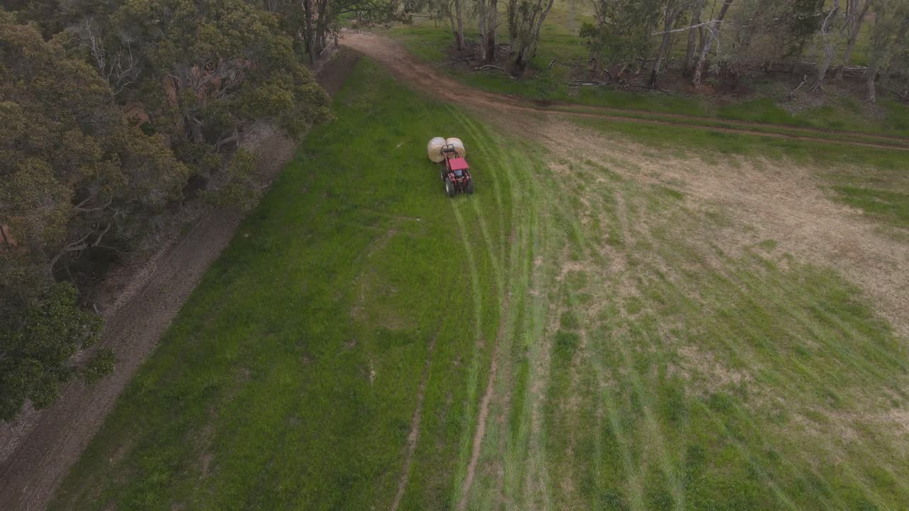 sobrevuelo aéreo que muestra un tractor rojo que toma un fardo de hierba de un campo agrícola en un día nublado
