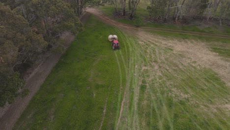 aerial flyover showing red tractor taking grass bale of agricultural field in cloudy day