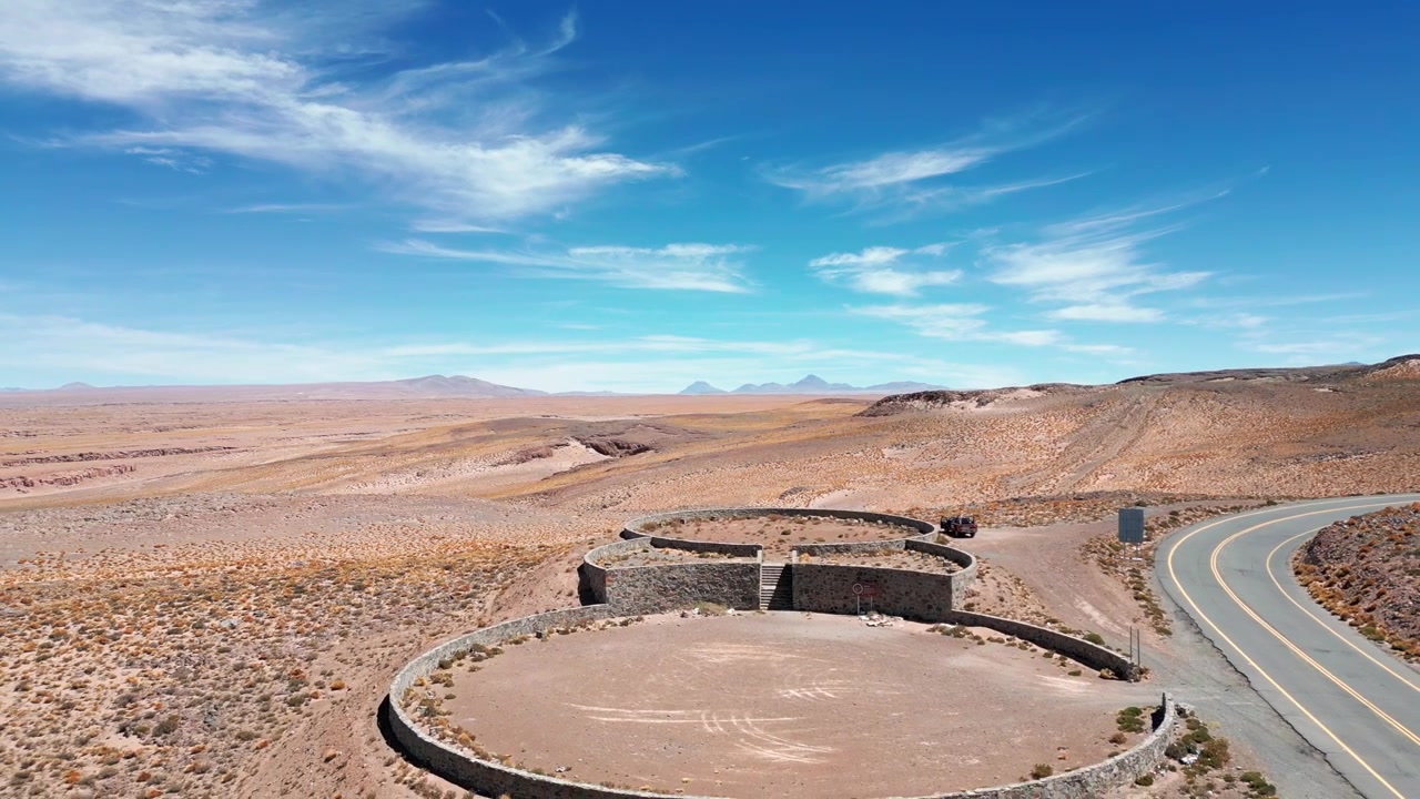 Backward reveal flight over circular stone lookout beside winding mountain road unveiling vast arid plateau with distant volcanic peaks. Paso Jama, Chile