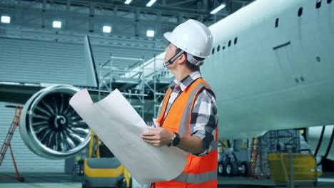 vista lateral de un ingeniero masculino asiático con casco de seguridad mirando el plano en sus manos y mirando a su alrededor mientras está de pie con el avión en el hangar