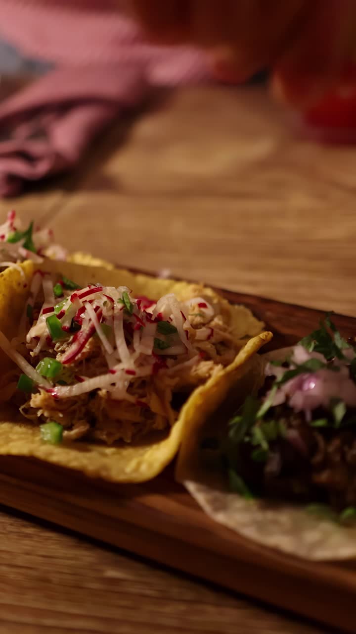 Una lenta panoramica della telecamera mostra tacos grigliati assortiti con condimenti freschi su un tagliere di legno, illuminati calorosamente in un accogliente ambiente da pranzo interno.