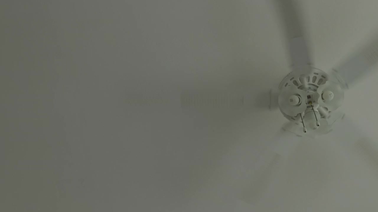 White ceiling fan spinning indoor room with lights on a clean white ceiling, creating cool airflow and breeze during summer. Handheld static shot of domestic ventilation appliance.