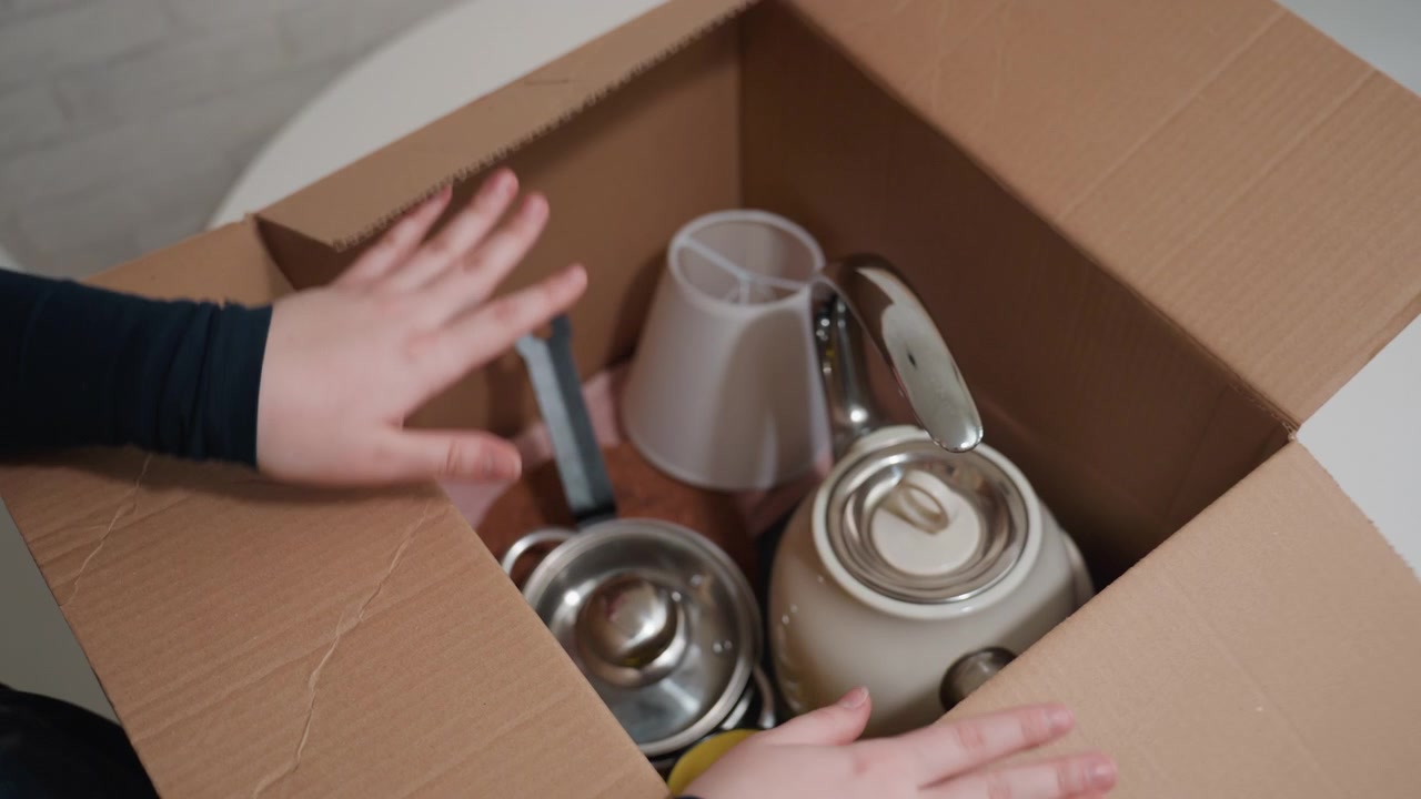 Packing kitchenware into cardboard box, hands placing kettle and mugs, closeup of cardboard flaps, careful arrangement, quiet moving day, small apartment table, new home preparation, fragile items