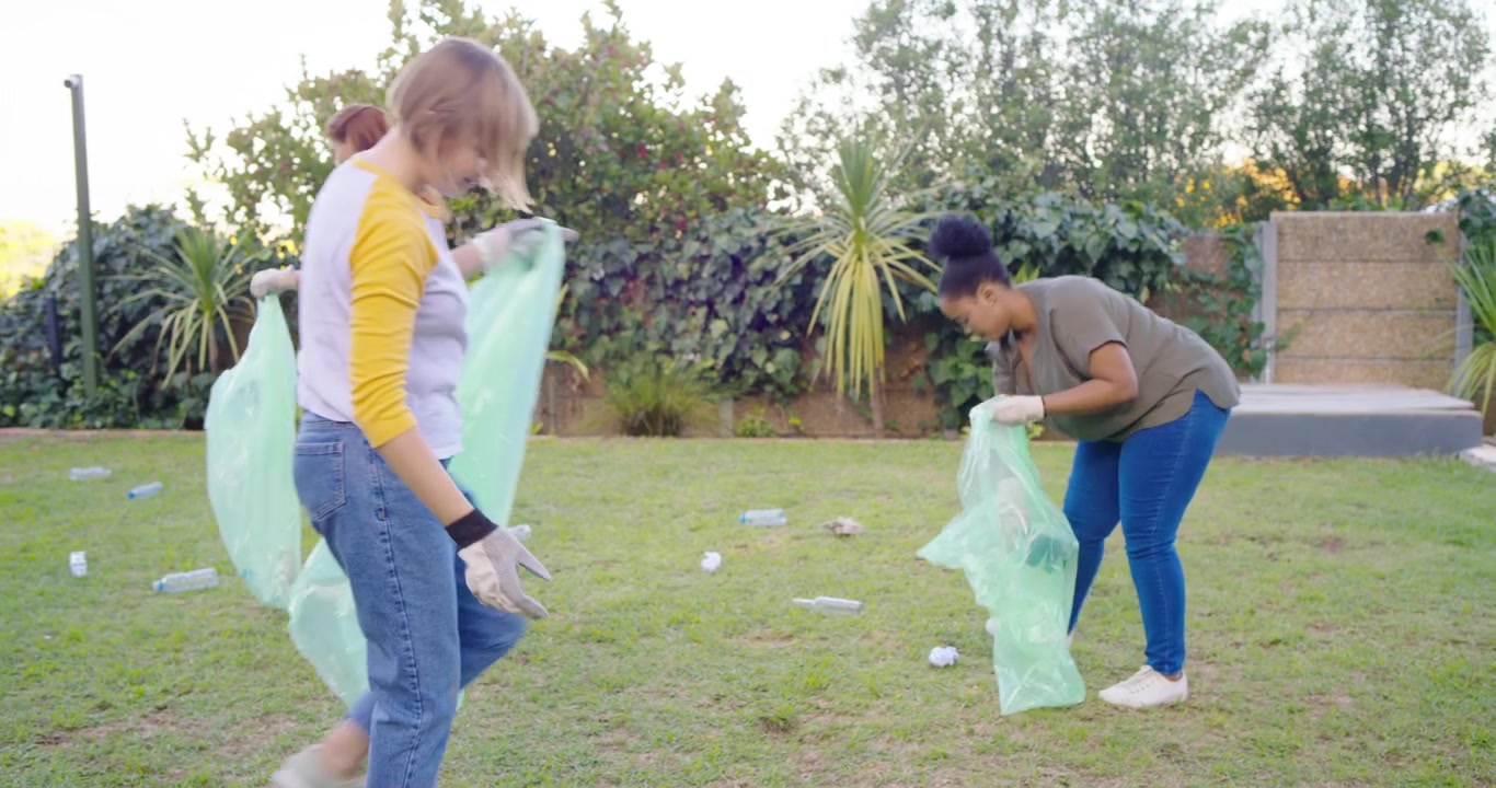 Diverse female friends seeing litter in backyard, donning gloves and filling green bags