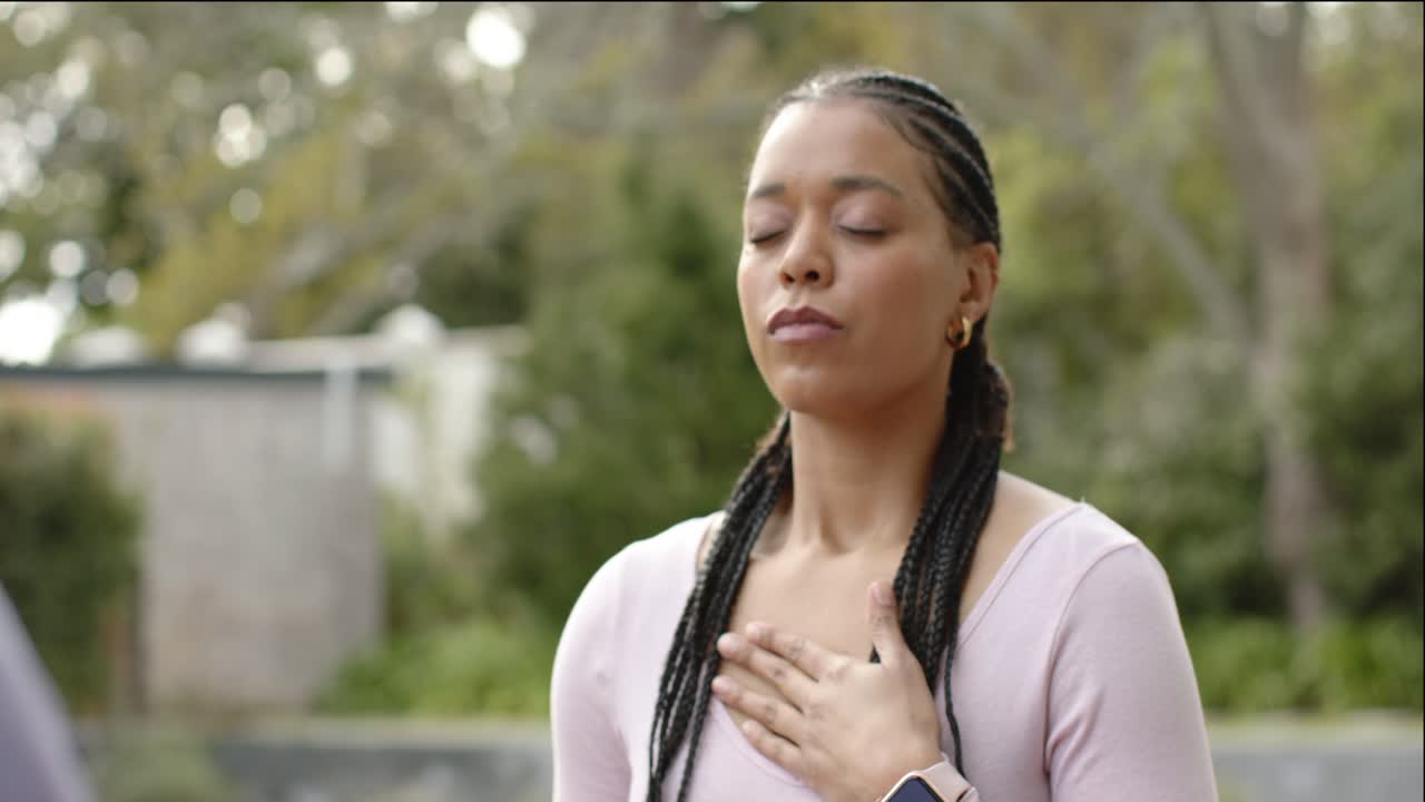 Woman placing hands on chest, breathing deeply to calm mind in garden, smartwatch visible