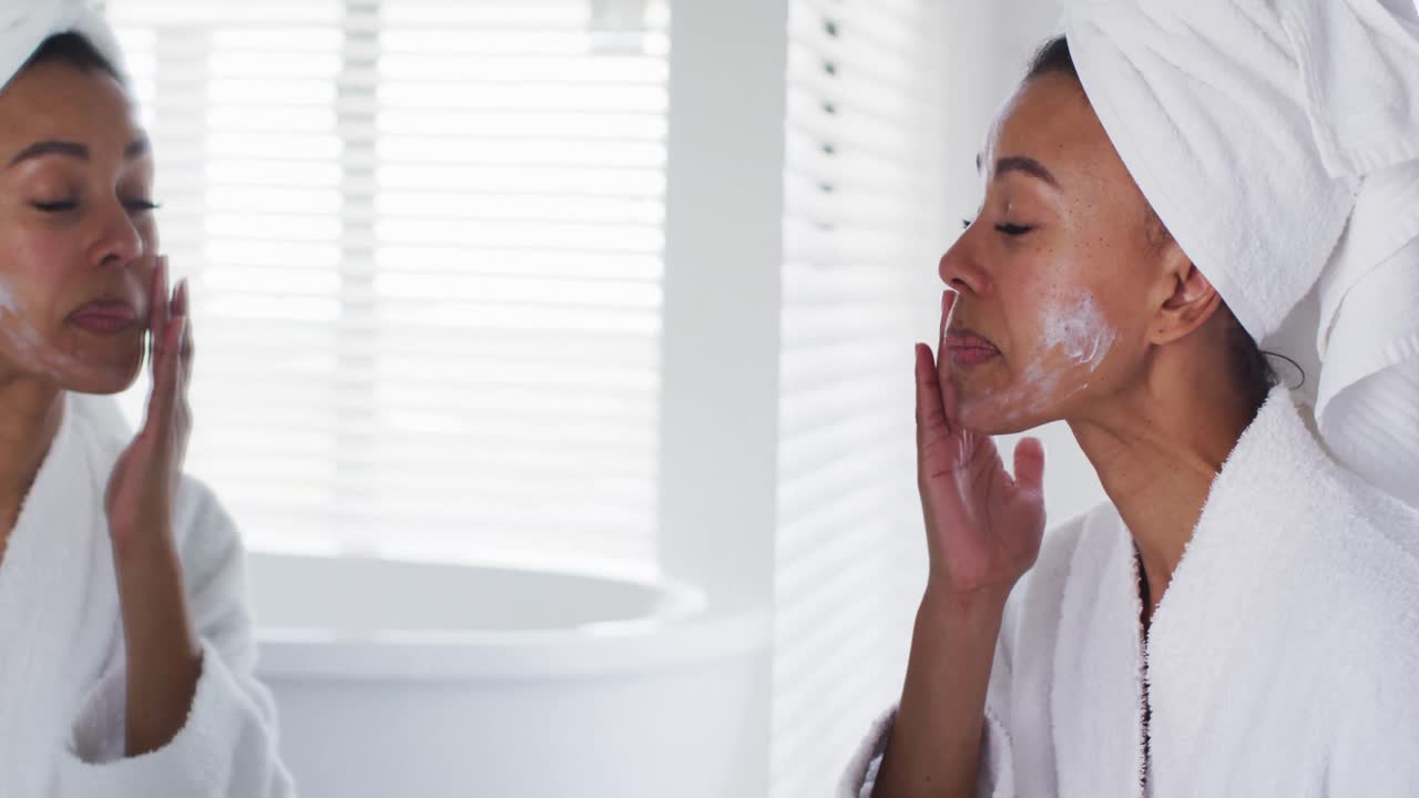 African american woman in bathrobe applying face cream looking in the mirror at bathroom