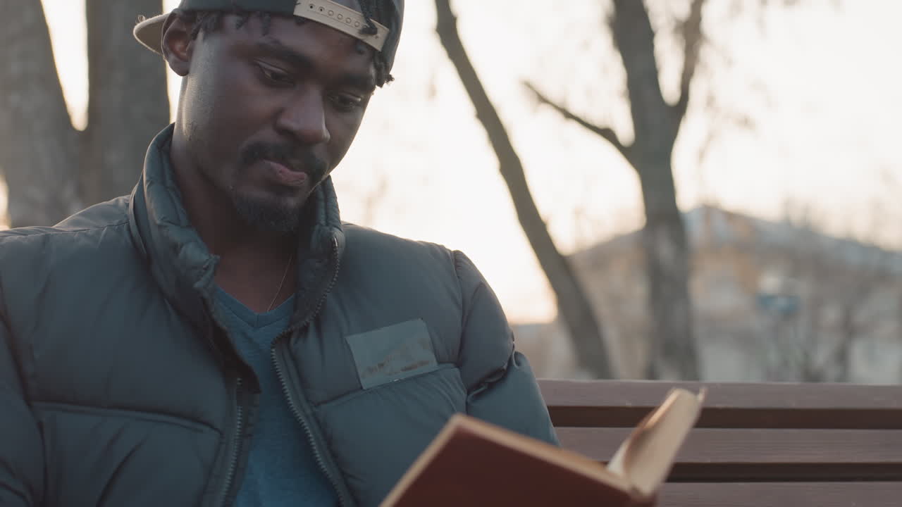 Young man in winter jacket and backward cap sitting on park bench reading hardcover book with deep focus, blurred background of trees and warm evening light
