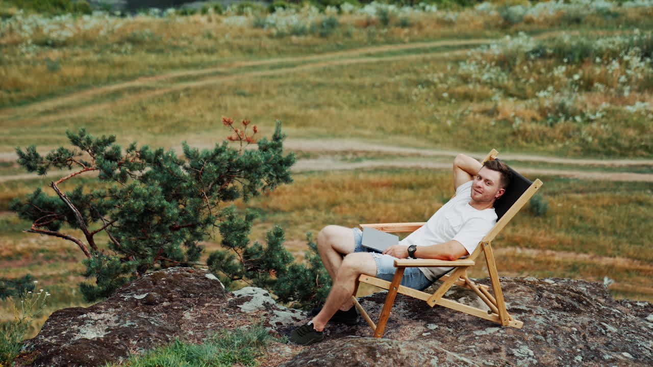 Relaxed Caucasian man having rest in the folding chair. Man sits on the rock looking at beautiful nature around. Freelancer with laptop working remote.
