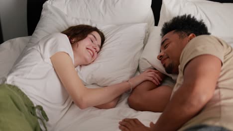 A-happy-young-adult-brunette-girl-with-a-bob-hairstyle-in-a-white-T-shirt-lies-on-the-bed-with-her-young-boyfriend-with-Black-brunette-skin-and-touches-his-hand-in-the-morning