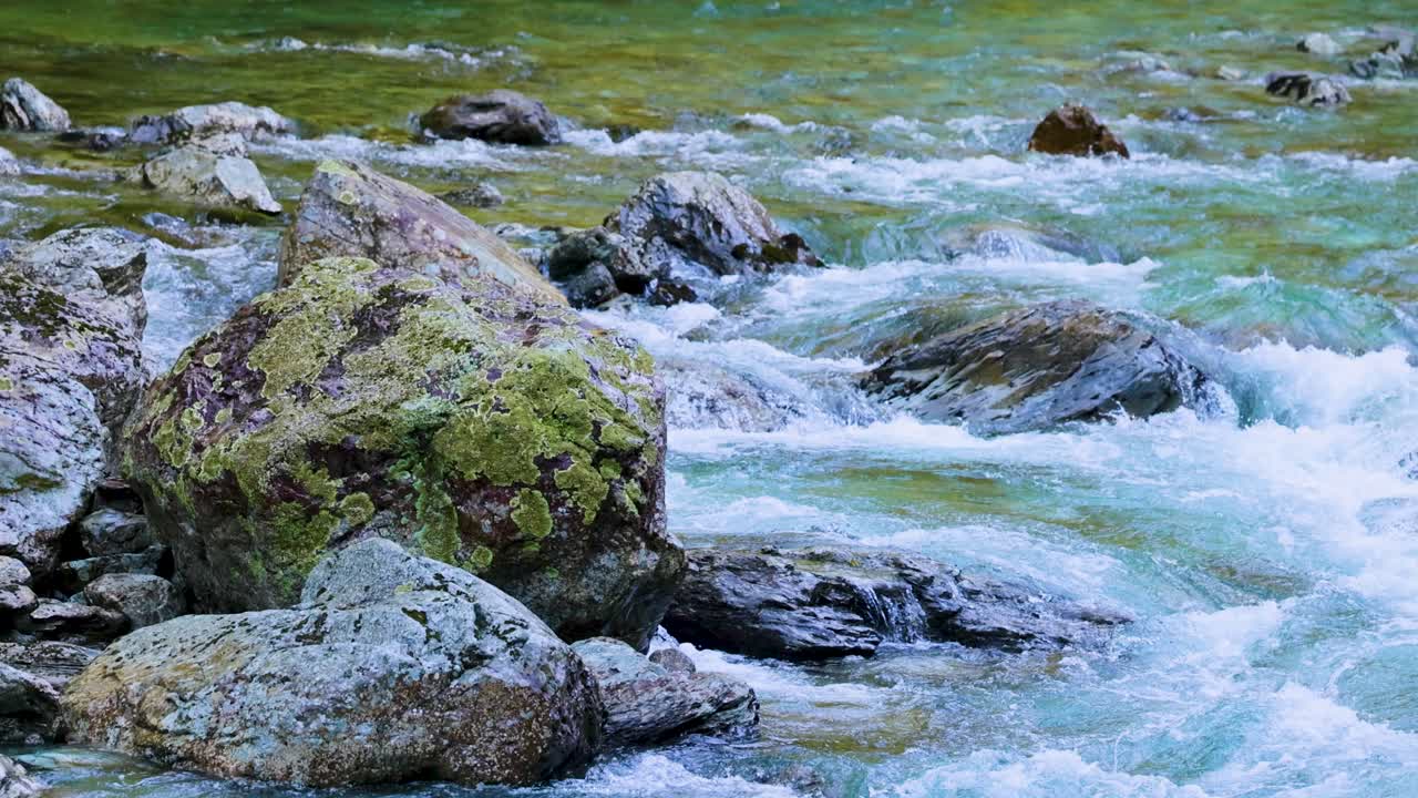 Fast-moving turquoise river water rushes over moss-covered rocks in bright, natural daylight