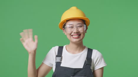 close up of asian woman worker wearing goggles and safety helmet smiling, waving hand, and saying bye while standing in the green screen background studio