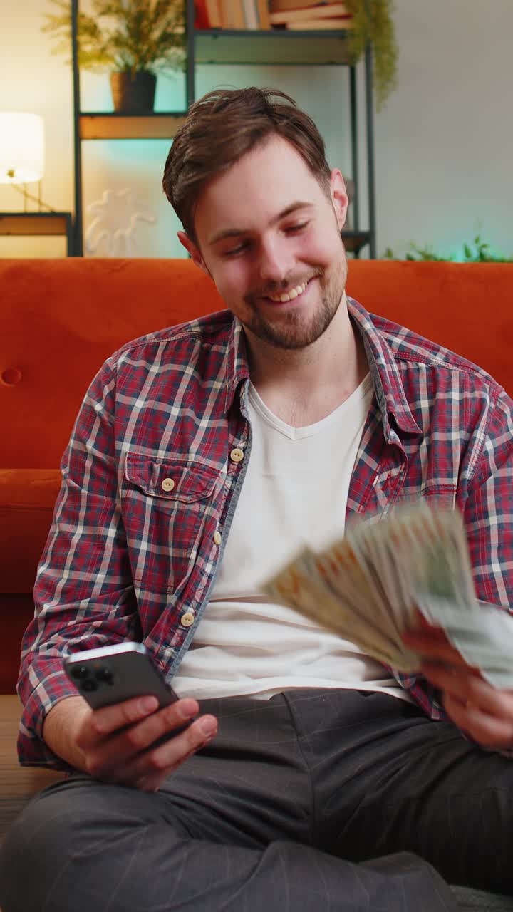 Successful happy rich man holds smartphone waving dollar bill money fan sitting in room at home