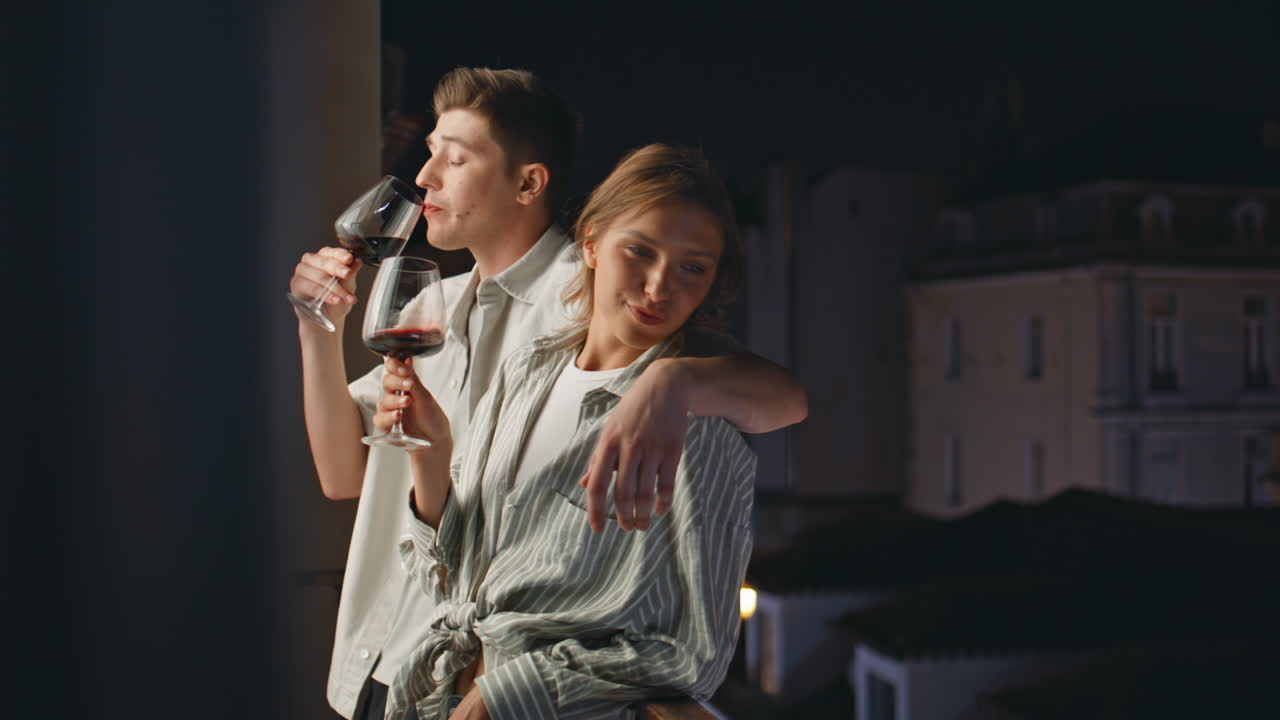 Smiling sweethearts drinking wine on balcony. Cheerful couple dating at night