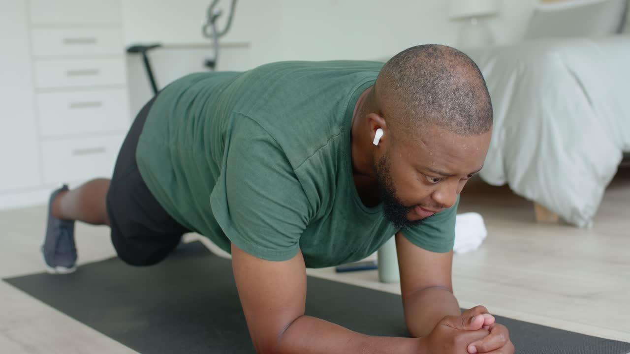 African American man starting forearm plank in bedroom, wearing green tee and earbuds, toning core