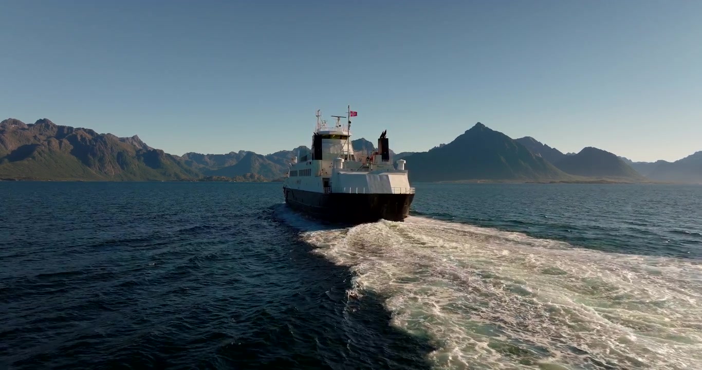 Drönare följer färja som kryssar över blått vatten mot berg från Melbu till Fiskebøl på Lofoten, Norge. Flygfoto, utrymme för text.