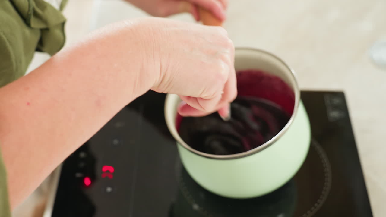 Cook wearing green sleeve stirs thick red porridge inside pastel green pot placed on electric stove while food simmers, steam rising gently in bright home kitchen during cooking process