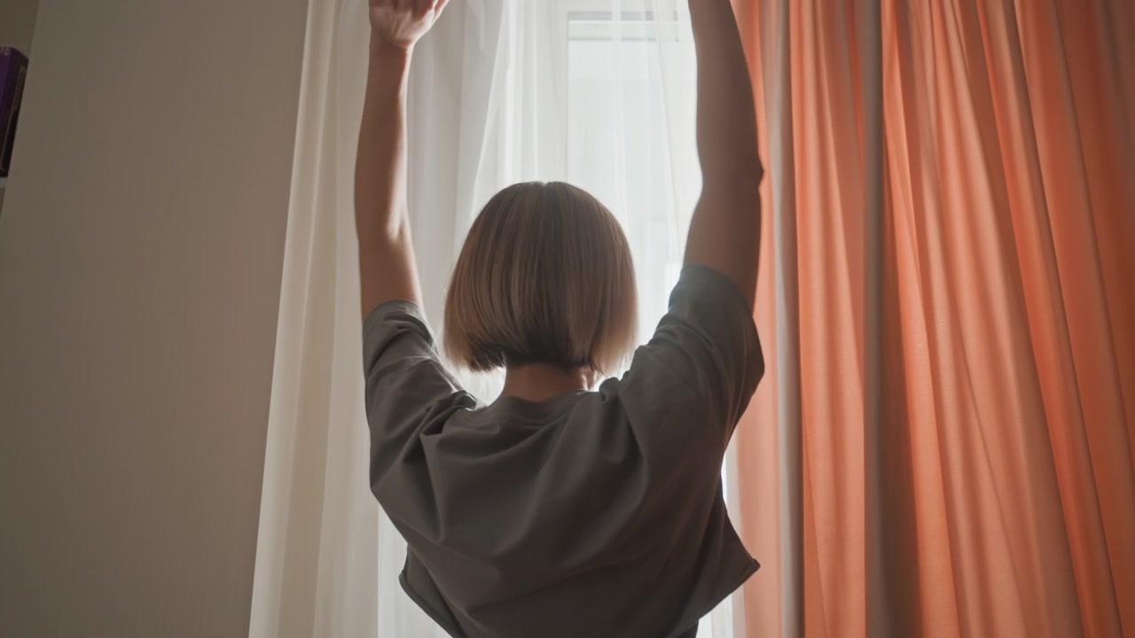 Frau öffnet korallenfarbene Vorhänge im Morgengrauen. Silhouetten-Sequenz in einem ruhigen Schlafzimmer, kleine blonde Frau steht im Schatten, zieht dicken Vorhang zurück, um sanfte Morgenstrahlen zu enthüllen, streckt die Arme nach oben, blickt