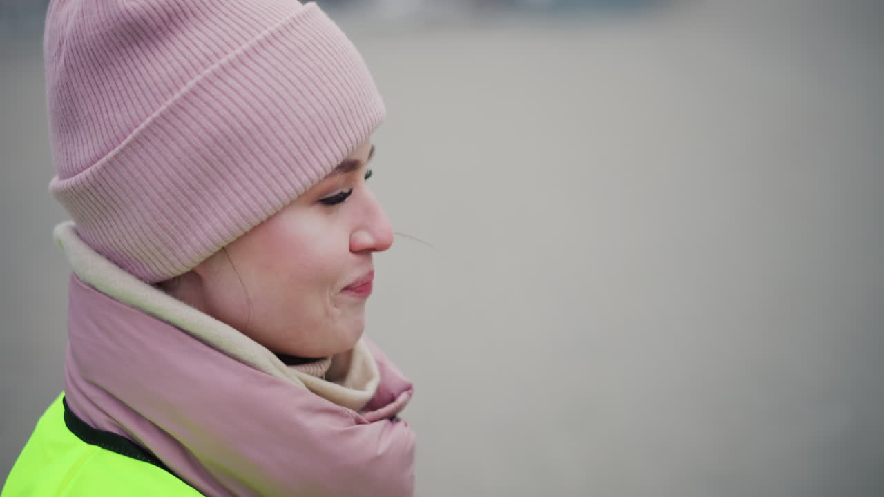 female worker wearing neon reflective safety vest, pink beanie, and warm scarf smiling while looking down, standing outdoors in cold weather, suggesting casual conversation, break time during outdoor work