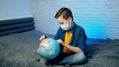 child in self isolation looking at globe learning about covid-19. boy in a medical mask with curious face thinking about the planet during covid lock down, quarantine and social distancing concept