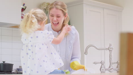 Mother-wearing-rubber-gloves-at-home-in-kitchen-with-young-daughter-having-fun-as-girl-puts-feet-in-sink-as-they-do-washing-up-at-sink--shot-in-slow-motion