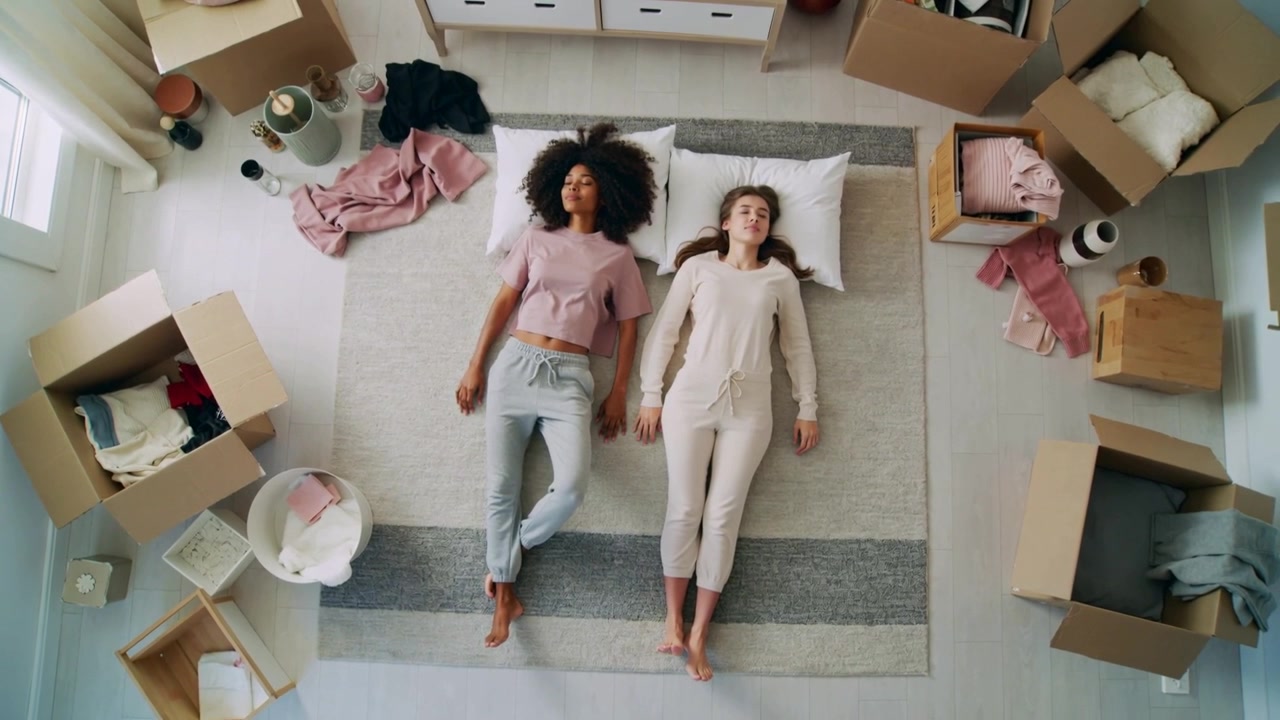 Pausing unpacking two women lying on rug wearing pink top beige set sharing glances near boxes