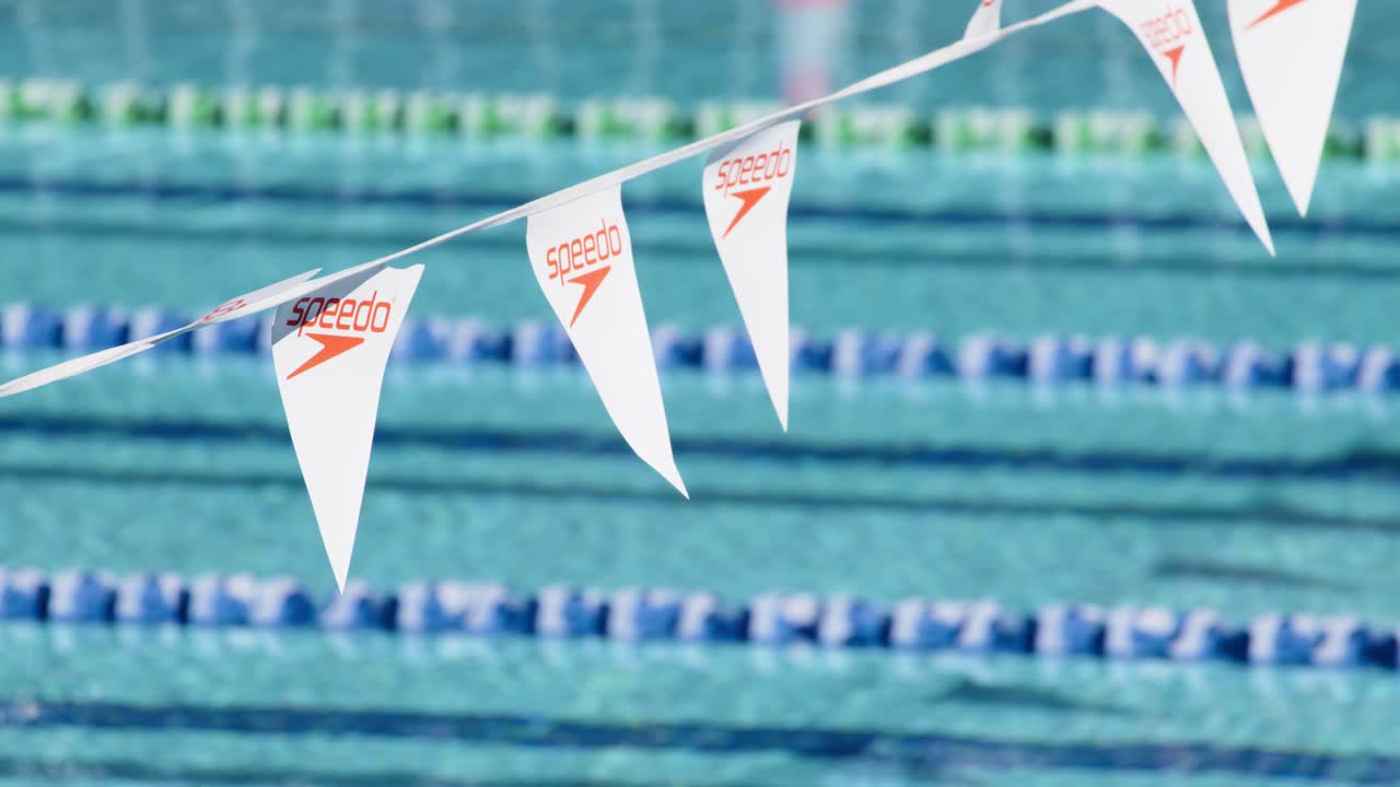 White triangular lane flags flutter above a clear outdoor swimming pool with blue lane dividers, captured in bright natural daylight with a shallow depth of field