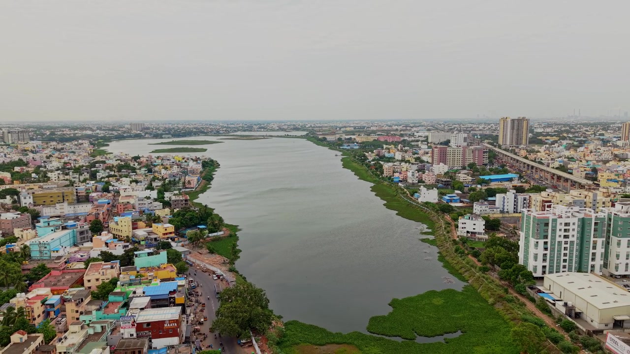 Luftfoto fra drone af bred flod, der løber gennem tæt bebygget byområde, med farverige huse, veje og højhusbygninger langs begge bredder i Chennai, Tamil Nadu, Indien