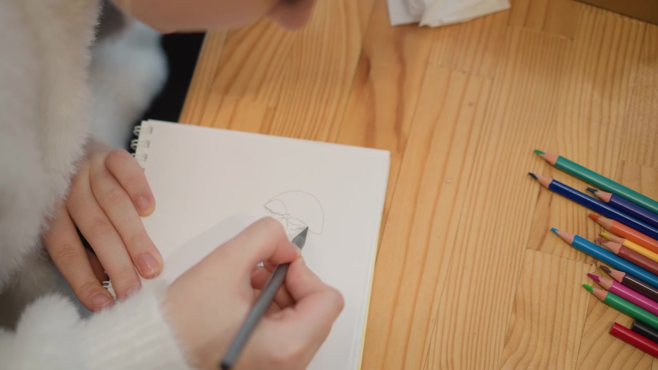 Overhead close-up shows hands sketching on blank notebook page with colored pencils on rustic wooden table, with visible drawing motion, writing activity, planning focus, and calm creative workspace