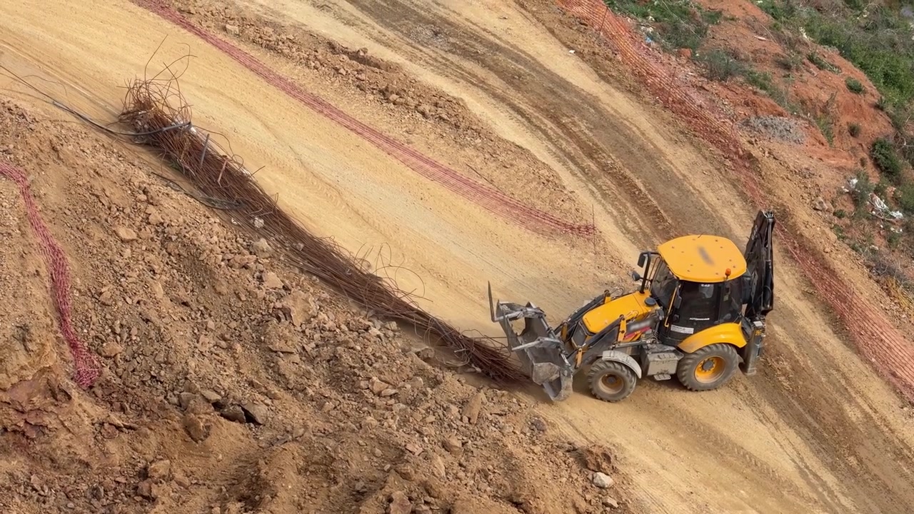Backhoe loader handling steel rebars and working on a steep slope at a construction site [cite: MM.jpg]. Infrastructure development and slope stabilization field operation