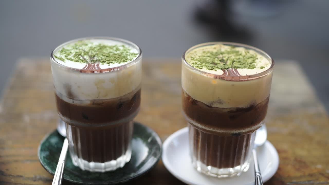 Üzeri üç kremsi köpükle süslenmiş iki mocha, sosluklarda cam bardaklarda katmanlı espresso ve süt, bulanık sokak arka planı açık hava kafe havası yaratıyor, sıcak palet ve yakın çekim kadrajlama.