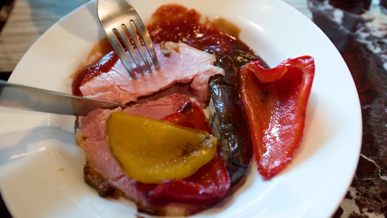 Una persona usa una forchetta e un coltello in acciaio inossidabile per tagliare prosciutto arrosto con peperoni grigliati.